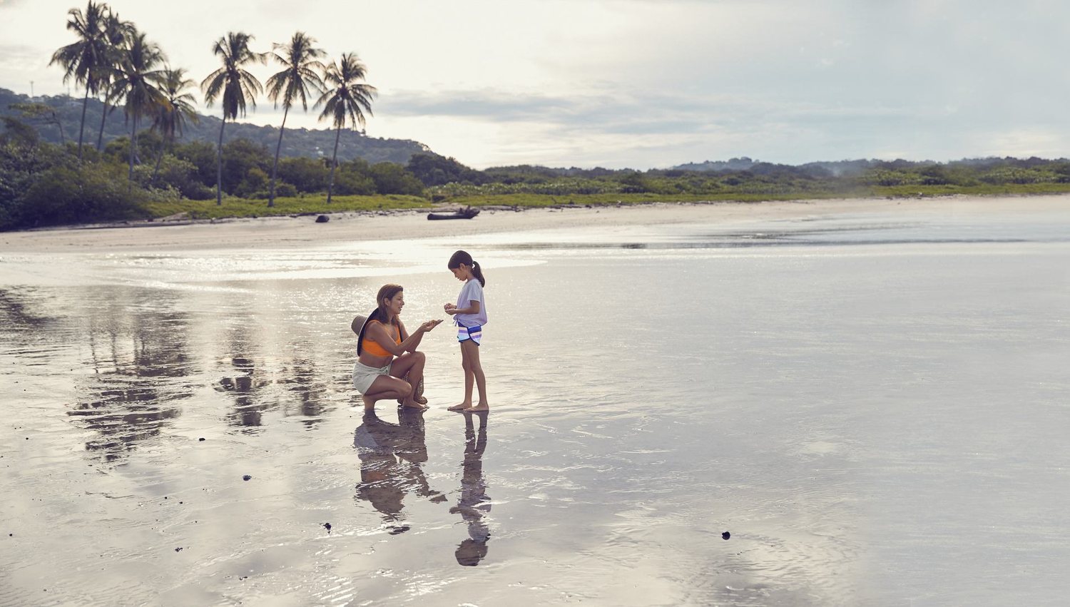 Carolina Sevilla crouching beside a child on a wide reflective beach at low tide, showing her something held in her hand